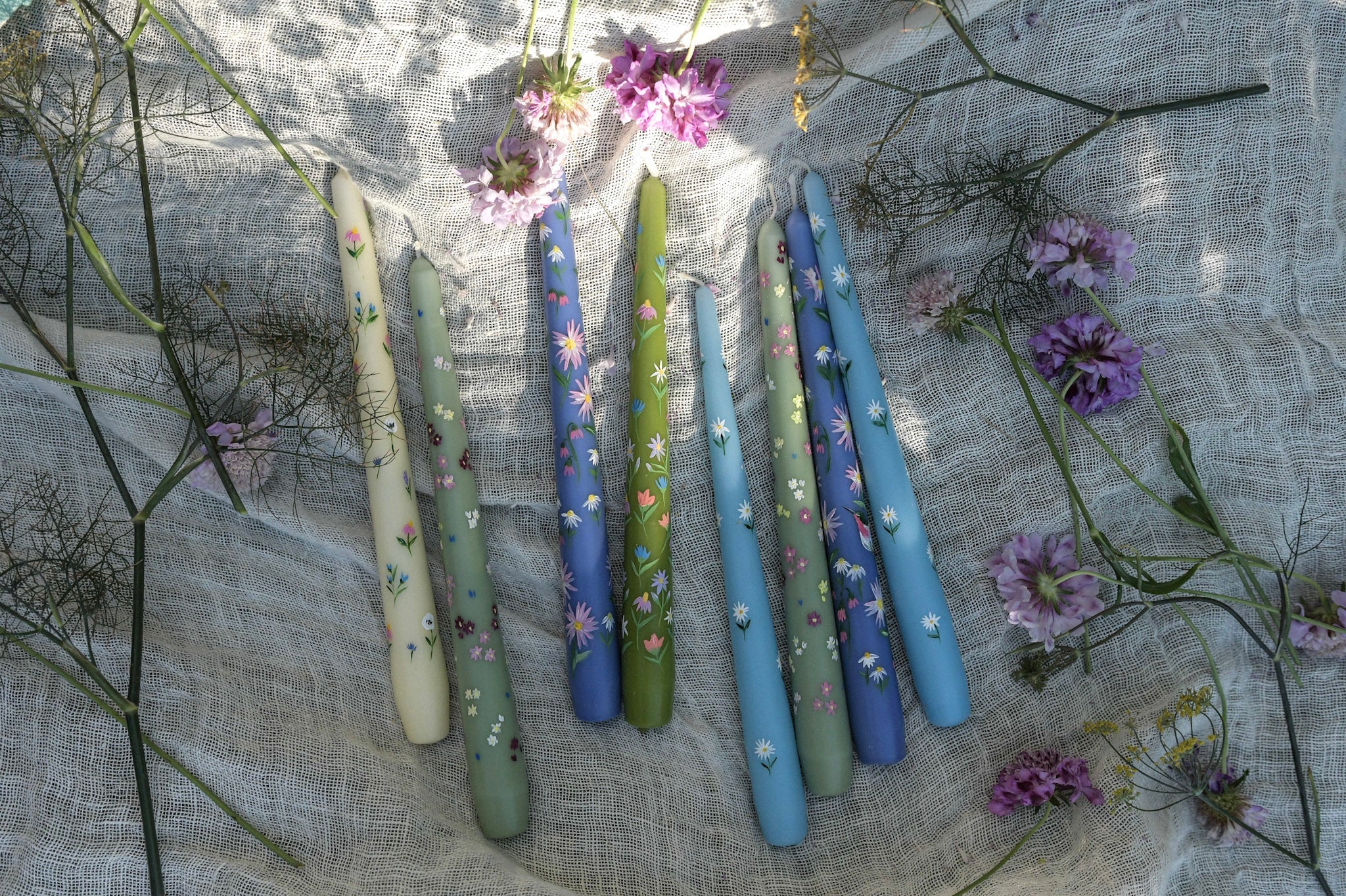 taper dinner candles are hand painted with floral designs. the candles are surrounded by flowers and a linen table cloth